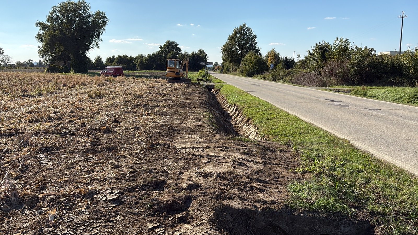 Terreno appena ripulito lungo una strada di campagna, con il fossato di scolo riaperto e un escavatore al lavoro sullo sfondo