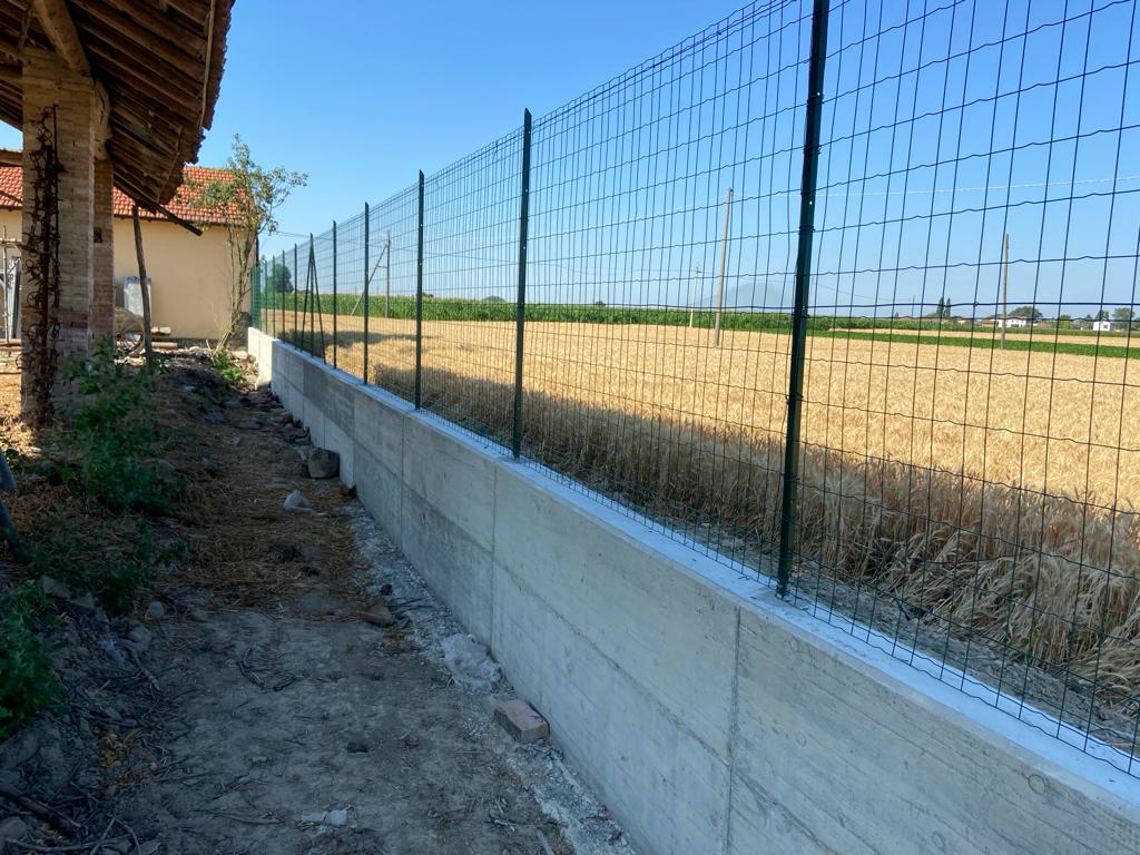 Muretto in calcestruzzo gettato lungo il confine di una proprieta', con recinzione metallica verde fissata sopra e campo coltivato a fianco