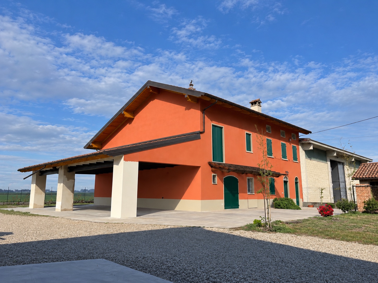 La stessa cascina a lavoro finito: intonaco arancio con zoccolatura chiara, serramenti verdi, tetto nuovo con travi in legno a vista, portico aperto sul fianco e piazzale sistemato